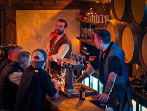 Intérieur d'une taverne médiévale-fantastique faiblement éclairée par des lanternes vacillantes, murs en roche noire, mobilier en bois sculpté sur mesure, et une grande fenêtre-écran laissant apercevoir un paysage fantastique animé.
