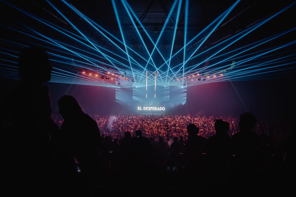 Vue d'une grande salle de concert plongée dans l'obscurité, traversée de faisceaux lumineux colorés et d'effets stroboscopiques, foule compacte de festivaliers déguisés pour Halloween dansant face à une scène.