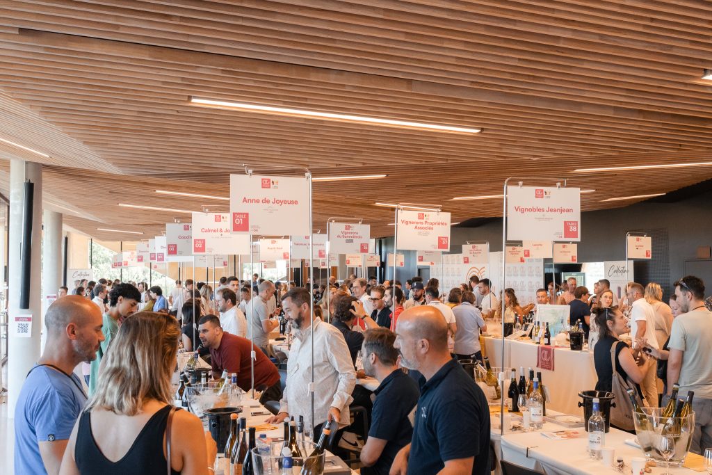Une soirée de dégustation de vin conviviale et chaleureuse dans un grand espace événementiel, visiteurs tenant des verres de vin près de stands de dégustation, ambiance animée et festive de fin d'après-midi automnale.
